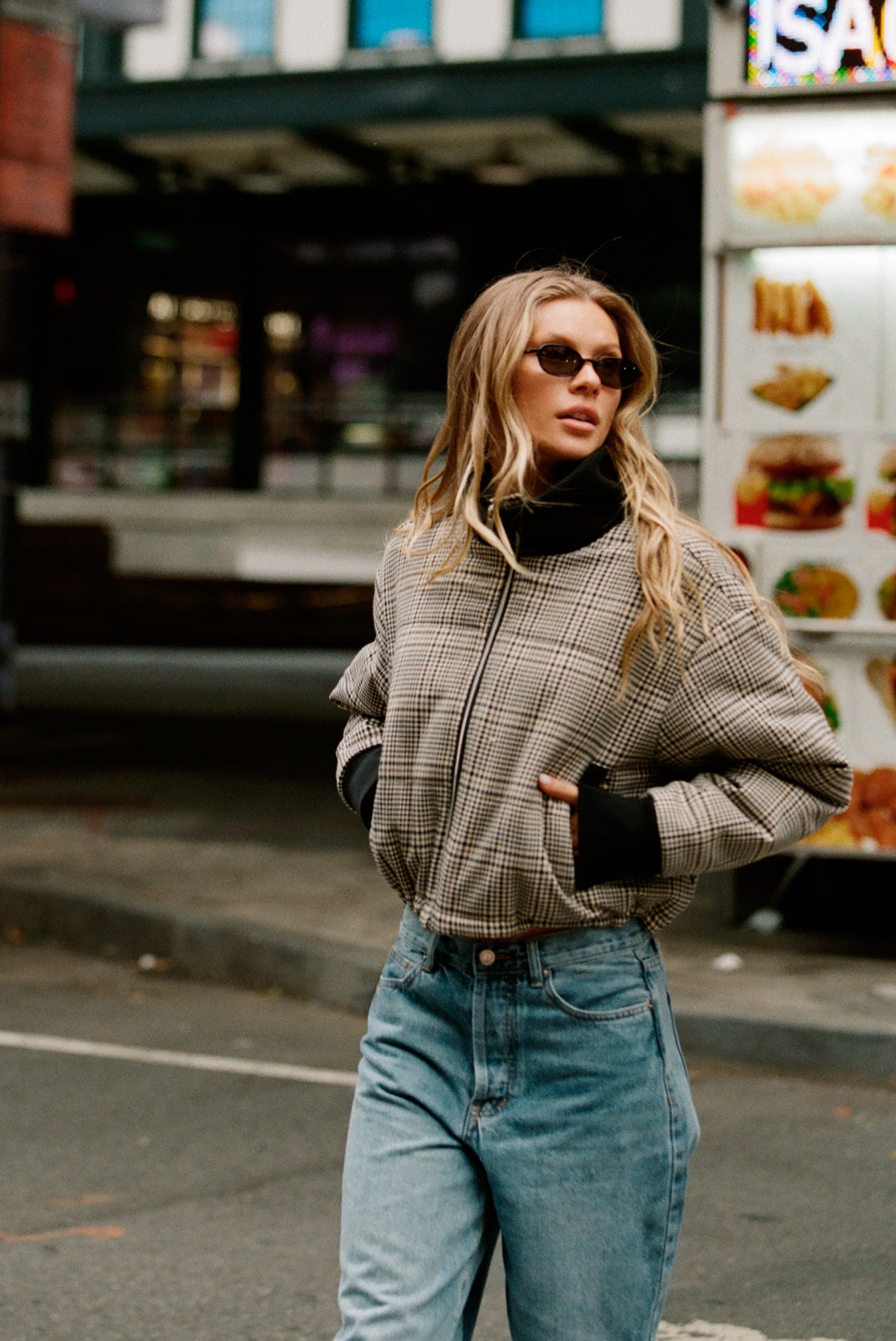 A woman wearing a beige and brown plaid zip-up jacket over a black turtleneck, paired with light blue jeans, standing on a city street.