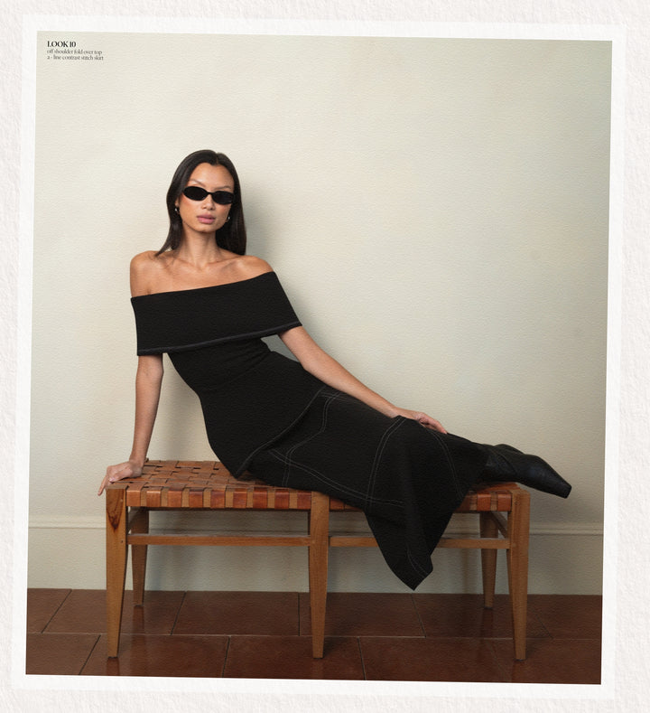 Woman in a black off-shoulder matching set sitting on a wooden bench against a plain wall.