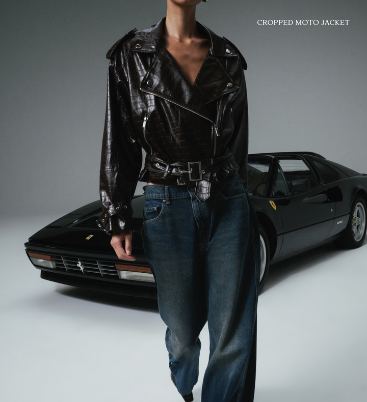 Person wearing a cropped moto jacket in front of a black sports car on a gray background