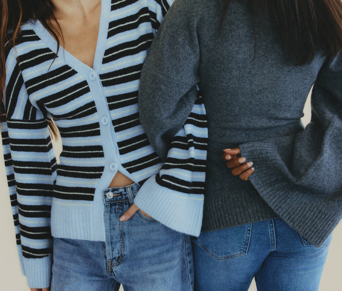 Two women wearing knit cardigans, one light blue with black horizontal stripes and one solid dark gray, paired with blue jeans.